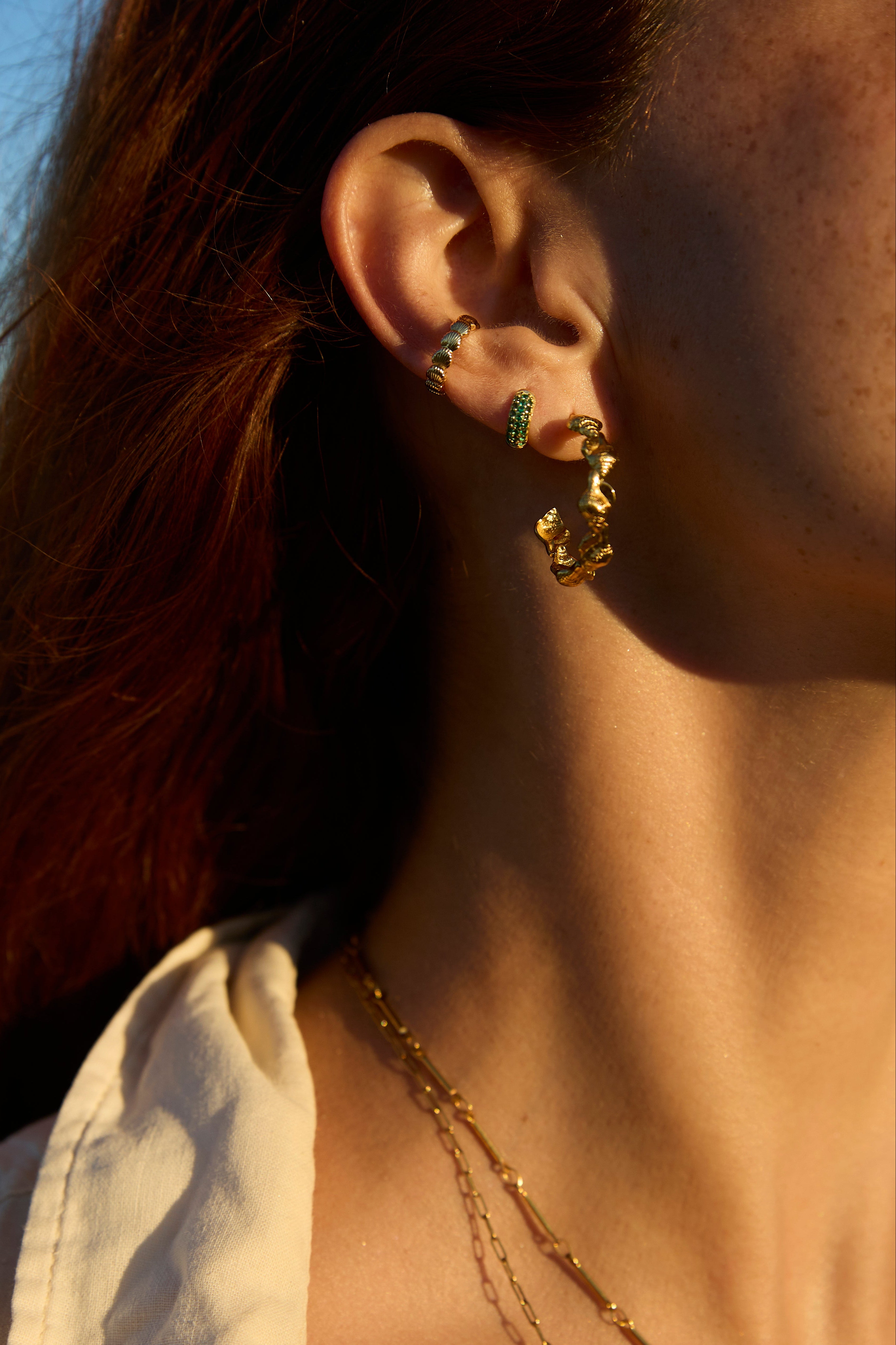 A close-up lifestyle shot of a model wearing gold hoop earrings with green crystal accents, layered with a delicate gold chain necklace.